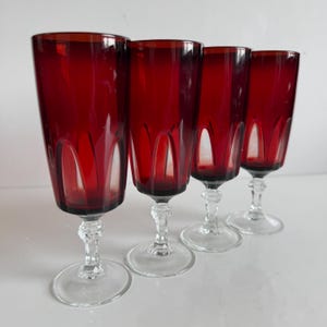 May include: Four ruby red stemmed glasses. The glasses have a tall, fluted design and clear, ornate stems and bases. The glasses are arranged in a row, showcasing their elegant shape and colour. Ideal for serving drinks.