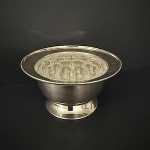May include: A silver flower frog with a clear glass insert. The frog sits in a round bowl with a beaded rim and a pedestal base. The glass insert has multiple holes for arranging flowers.
