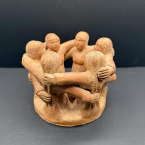 May include: A terracotta sculpture featuring a circular group of six figures standing in a ring, each with arms linked around the shoulders of the others. The figures have textured hair and simple facial features, all rendered in a matte clay finish.