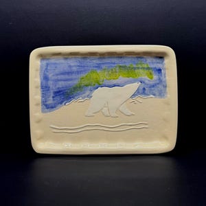 May include: A rectangular cream-colored ceramic tray featuring a raised white polar bear relief in the center. The background depicts a painted blue sky with a vibrant green aurora borealis effect. The tray has a textured border with small indentations.
