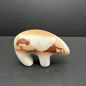 Könnte beinhalten: Keramik-Bärenfigur in Beige und Weiß. Der Bär ist mit einer gemalten Landschaftsszene mit einem Baum, Bergen und einer Behausung verziert. Der Bär hat eine glatte, abgerundete Form und einen flachen Sockel.