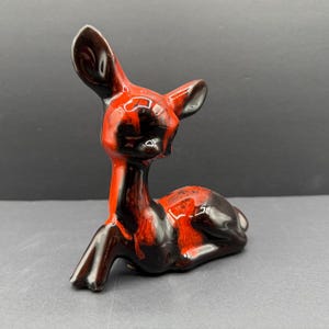 May include: A ceramic deer figurine in a reclining position, featuring a high-gloss glaze with a marbled pattern of deep black and vibrant orange-red colors. The deer has large, upright ears and a smooth, reflective surface finish throughout.