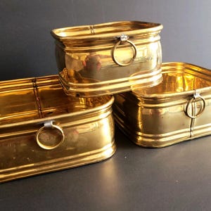 Peut inclure: Trois récipients rectangulaires en laiton aux coins arrondis et aux surfaces dorées polies. Chaque récipient présente une poignée en anneau métallique fixée au centre de la face avant, avec un design nervuré horizontal sur la base et le bord.