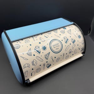 May include: A light blue bread box with a roll-top lid. The front panel is cream-colored and features a repeating pattern of dark blue line-art illustrations of pastries, cakes, and cutlery, with a central circular logo reading BAKERY DELICIOUS FRESH.