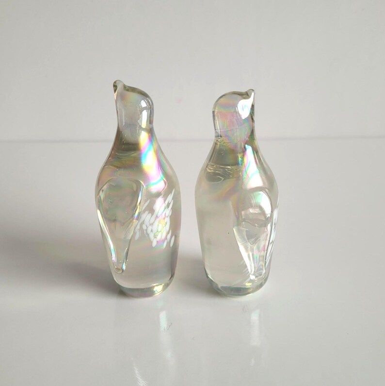 May include: Two iridescent glass bud vases with a pearlescent sheen. The vases have a unique, curved shape with a slender neck and a rounded body. The glass reflects a rainbow of colors, adding a touch of elegance and visual interest. These vases are perfect for displaying small flowers or stems.