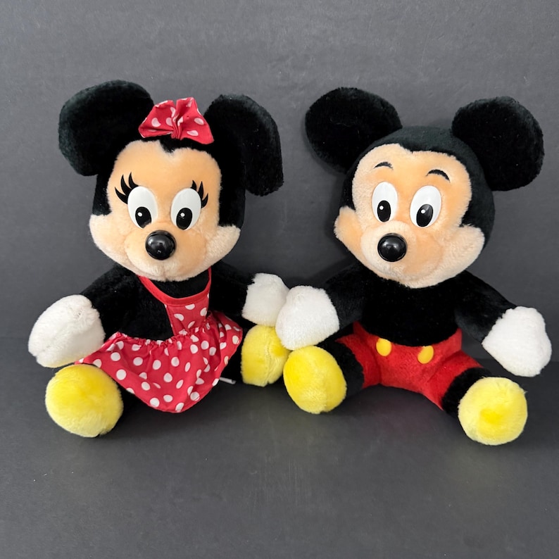 May include: Two plush Disney character dolls, Mickey Mouse and Minnie Mouse, sit side-by-side. Mickey wears red shorts and yellow shoes. Minnie wears a red polka-dot dress and a red bow. Both have black fur and tan faces.