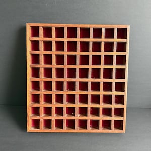 May include: A rectangular wooden display case featuring a grid of small square compartments. The frame and dividers are constructed from medium-toned brown wood, and each individual cubby is lined with a deep red velvet fabric backing.
