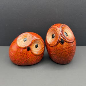 May include: Two ceramic owl figurines featuring a vibrant orange and red mottled glaze. Each owl has large, circular, textured facial discs surrounding black eyes and a small black beak. One owl is rounded and squat, while the other is taller.