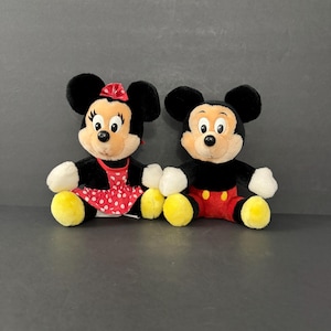 May include: Two plush Disney characters, Minnie Mouse and Mickey Mouse, sit side-by-side. Minnie wears a red polka-dot dress and bow. Mickey wears red shorts and yellow shoes. Both have black ears and smiling faces.