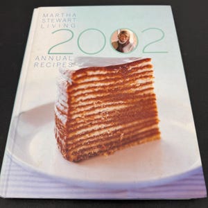May include: A cookbook titled "Martha Stewart Living 2002 Annual Recipes" with a slice of layered cake on the cover. The cover is light blue with the title in white and green. A photo of Martha Stewart is in the upper right corner.