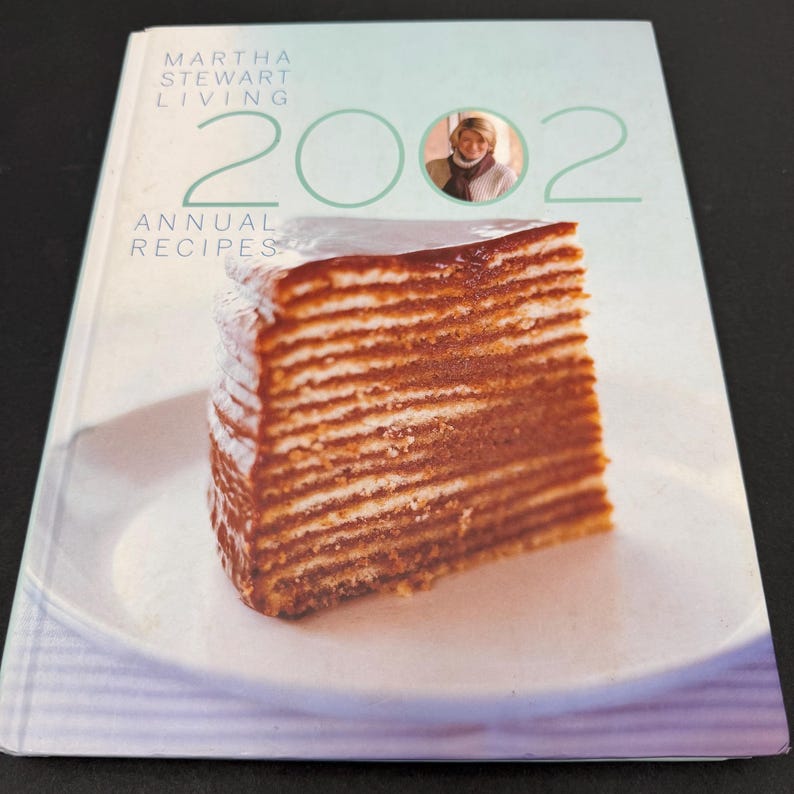May include: A cookbook titled "Martha Stewart Living 2002 Annual Recipes" with a slice of layered cake on the cover. The cover is light blue with the title in white and green. A photo of Martha Stewart is in the upper right corner.