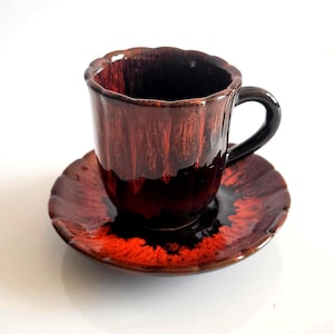 May include: A ceramic coffee cup and saucer set. The cup and saucer are glazed in shades of brown and red, with a glossy finish. The cup has a scalloped rim and a black handle. The saucer has a matching scalloped edge.