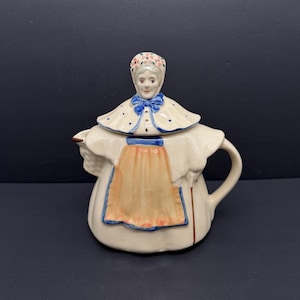 Könnte beinhalten: Eine keramische Teekanne in Form einer Frau, mit Blumenhaube und blauer Schleife. Die Teekanne hat einen cremefarbenen Sockel mit einer pfirsichfarbenen Schürze und blauen Verzierungen. Der Deckel ist mit blauen Punkten verziert.