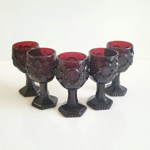 May include: Five ruby red wine glasses with ornate detailing. The glasses have a dark stem and a patterned bowl. They are displayed on a white surface, highlighting their rich colour and design. Each glass is approximately 15 cm tall.