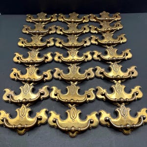 May include: A collection of eighteen brass-toned metal drawer pulls arranged in six rows of three. Each pull features a decorative backplate with a central vertical motif and a curved, swinging handle attached to two mounting points on the plate.