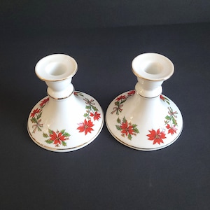 May include: A pair of white ceramic candle holders with a gold trim and a festive poinsettia design. Each base features red poinsettias, green leaves, and ribbons. These candle holders are a classic Christmas decoration.