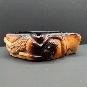 May include: A carved brown and dark amber-toned ashtray featuring a relief sculpture of a reclining figure with textured details. The object has a smooth, polished finish with deep brown accents and a central depression for holding items.