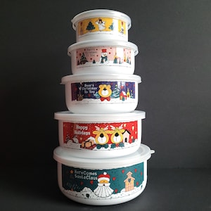 Könnte beinhalten: Ein Stapel aus fünf weißen, runden Vorratsbehältern mit transparenten Deckeln, die jeweils weihnachtliche Illustrationen und Texte wie Merry Christmas To You, Bear's Christmas To You, Happy Holidays und Here Comes Santa Claus aufweisen.