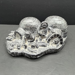 May include: A dark grey stone carving depicting two igloos, two figures in hooded parkas, and a small animal. The sculpture features textured block patterns on the igloo domes and detailed carvings of the figures and wildlife on the base.