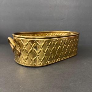 Può includere: Una fioriera ovale color oro con un motivo a rombi e due manici. La fioriera è in metallo e ha una finitura lucida. I manici sono curvi e attaccati ai lati.