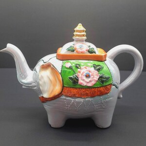 Puede incluir: Una tetera de cerámica con forma de elefante, cuerpo gris, con detalles en naranja y verde y un diseño floral. La tetera tiene un pico curvo, un asa redondeada y una tapa decorativa con un adorno dorado. La tetera mide unos 20 cm de largo.