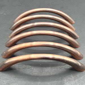 May include: A set of six arched cabinet pulls with a brushed copper finish. Each handle features a smooth, curved design with a metallic texture, arranged in a parallel row to showcase the consistent bronze-toned colour and rounded shape.