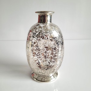 May include: Silver vase with a floral design. The vase has a rounded body, a narrow neck, and a flared base. The floral pattern is detailed and covers the entire surface. The vase is likely made of glass or metal, and is a decorative item.