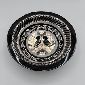 May include: A black and white decorative ceramic bowl. The bowl features a central design of two black birds perched on a floral pattern. The rim has a detailed, repeating pattern. The bowl is round and has a glossy finish.