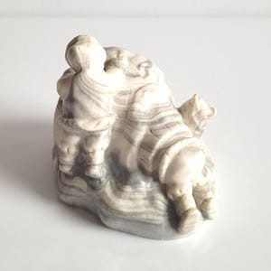 Op de afbeelding: Een gebeeldhouwde sculptuur met twee figuren en een ijsbeer. De sculptuur is gemaakt van een lichtgekleurde steen met grijze adering. De figuren zijn gekleed in traditionele kleding. De ijsbeer bevindt zich aan de rechterkant van de sculptuur.