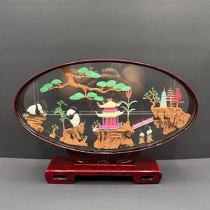 May include: An oval-shaped, framed diorama featuring a miniature Asian landscape. The scene includes pandas, pagodas, trees, and a bridge, all set against a black background. The frame and base are a deep red color.