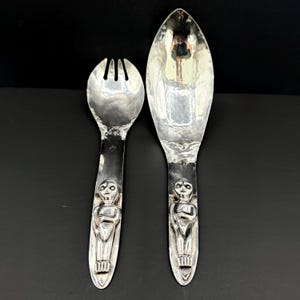Peut inclure: Une paire de couverts à salade en métal argenté poli, comprenant une fourchette à trois dents et une grande cuillère. Chaque manche présente un relief stylisé représentant une figure humaine intégrée au métal, avec des cuillerons lisses.