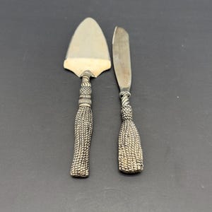 May include: Silver-toned cake server and butter knife set. The server has a triangular blade, while the knife has a rounded blade. Both have intricately designed handles with a woven texture, adding a touch of elegance to the serving set.