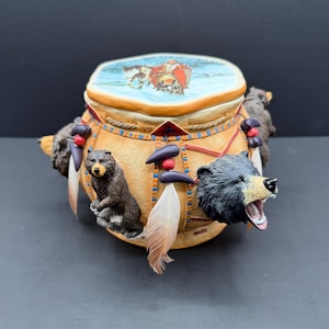 Può includere: Un barattolo in ceramica color cuoio con un coperchio raffigurante una scena dipinta con una figura in rosso. Il barattolo è decorato con figure di orsi tridimensionali, piume pendenti e perline scure. Un orso è in piedi, l'altro ha la bocca aperta.