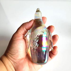 May include: Iridescent glass penguin figurine with etched floral design. The penguin has a clear body with rainbow hues and small wings. The figurine is held in a hand against a white background.