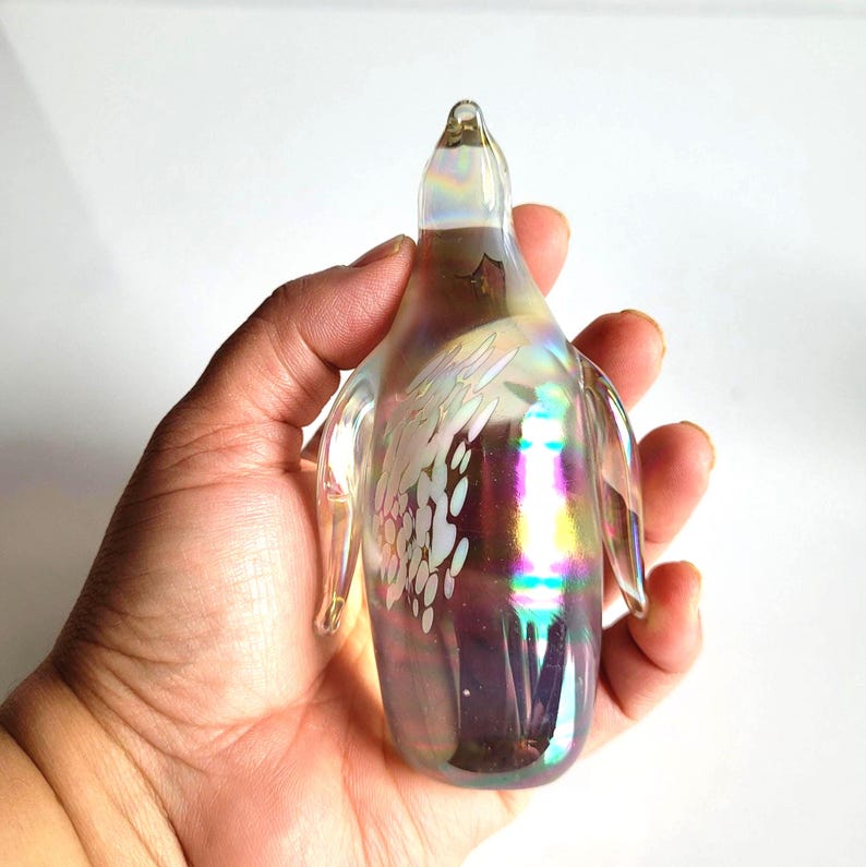 May include: Iridescent glass penguin figurine with etched floral design. The penguin has a clear body with rainbow hues and small wings. The figurine is held in a hand against a white background.