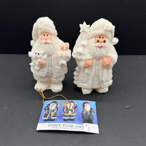Peut inclure: Deux figurines de Père Noël en céramique blanche non peinte avec des détails texturés. L'une tient un petit ours et un cadeau, l'autre un sapin de Noël. Inclut une carte d'instructions en papier marquée PAINT-YOUR-OWN Design Works Inc. Bohemia, NY.