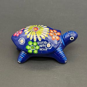 May include: A hand-painted, blue ceramic turtle figurine. The shell is decorated with colorful floral designs in yellow, orange, and green. The text "Riviera Maya Mexico" is written on the shell. The turtle has blue legs and head.