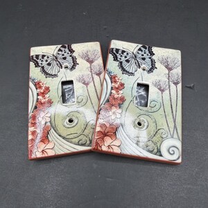 May include: Two rectangular light switch plates featuring a printed design of a black and white butterfly, thin brown floral stems, and clusters of small orange flowers against a pale green and cream background with swirling line patterns.