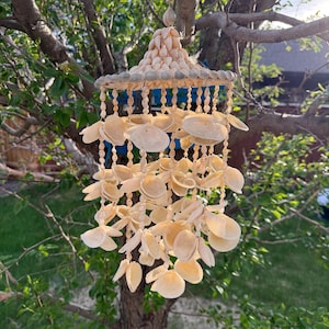 May include: A hanging mobile constructed from numerous off-white seashells. The structure features a conical top covered in small shells, with multiple strands of circular, flat shells suspended from a circular frame, creating a layered design.