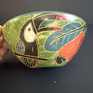 May include: An oval-shaped, hand-painted bowl with a toucan design. The bowl has a green background with a black toucan, and colourful leaves and flowers. The toucan has a white beak and is perched on a branch.