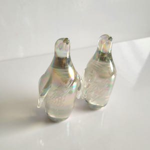 May include: Two iridescent glass penguin figurines. The penguins have a pearlescent sheen with hints of yellow, green, and blue. They have small wings and are standing upright. The glass is clear, allowing light to pass through.