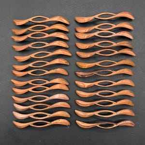May include: A collection of twenty-eight copper-toned metal cabinet pulls arranged in two vertical columns. Each pull features a slender, elongated, and curved design with a central open oval void, showcasing a smooth, metallic finish throughout.