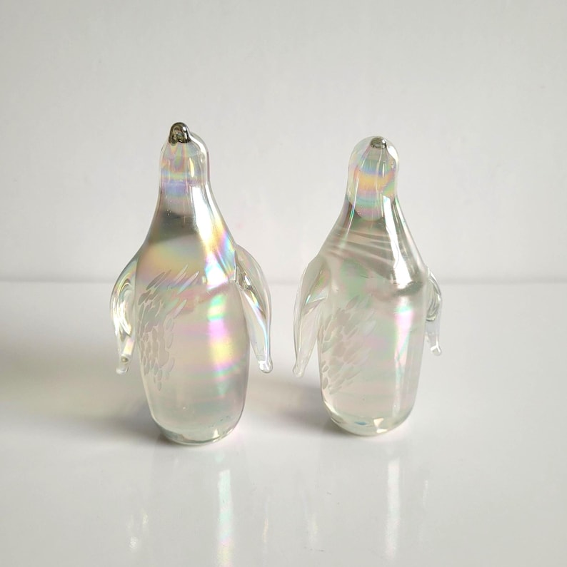 May include: Two iridescent glass penguin figurines. The penguins have a pearlescent finish with subtle rainbow hues. Each penguin has small, clear wings and a slightly pointed beak. The figurines are displayed on a white surface.