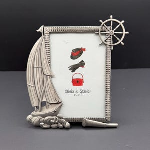 May include: Silver-coloured metal picture frame with a nautical theme. The frame features a sailboat, ship's wheel, and a spyglass. The picture inside shows a hat, gloves, and a purse. The text "Olivia & Gracie" and "4" x 6" is visible.