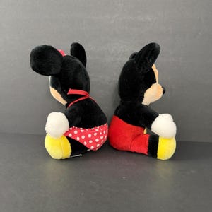May include: Two plush Disney character dolls, Mickey and Minnie Mouse, sit side-by-side. Mickey wears red shorts, while Minnie wears a red polka-dot skirt and a red bow. Both have black fur, white gloves, and yellow shoes.