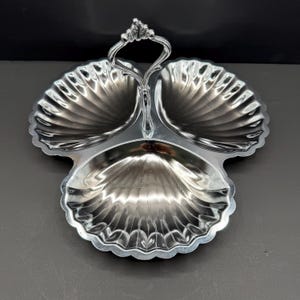 May include: A silver-toned metal serving dish featuring three distinct shell-shaped compartments arranged in a clover pattern. A central vertical handle with an ornate, decorative top loop connects the three sections, providing a grip for carrying.