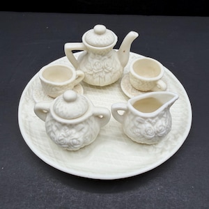 May include: A miniature off-white tea set featuring a teapot with a lid, a sugar bowl with a lid, a creamer, and two teacups on saucers. Each piece is decorated with a raised floral rose pattern and a textured lattice design on a round tray.