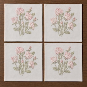 Puede incluir: Cuatro posavasos cuadrados de lino blanco, cada uno bordado con un ramo de rosas rosas y hojas verdes. Los posavasos están dispuestos sobre una superficie de madera oscura.