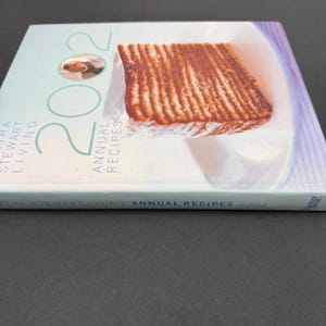 May include: A cookbook titled "Martha Stewart Living Annual Recipes 2002" with a light blue cover. The cover features a photo of a cooked dish and the number "2002" in large green numerals. The book's spine is visible.