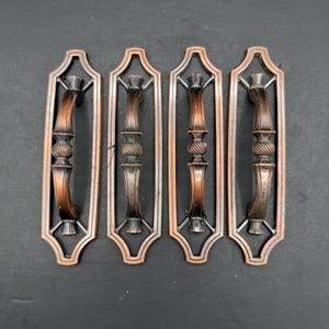 May include: Four matching drawer pulls with a copper-toned finish. Each pull features a central vertical handle with a textured, spiraled middle section, mounted within a rectangular, open-frame backplate with rounded, decorative corners.